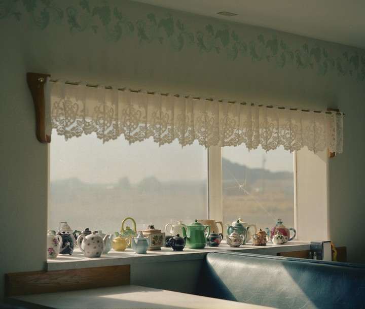 A collection of decorative teapots sits on a windowsill with a lace curtain above them.