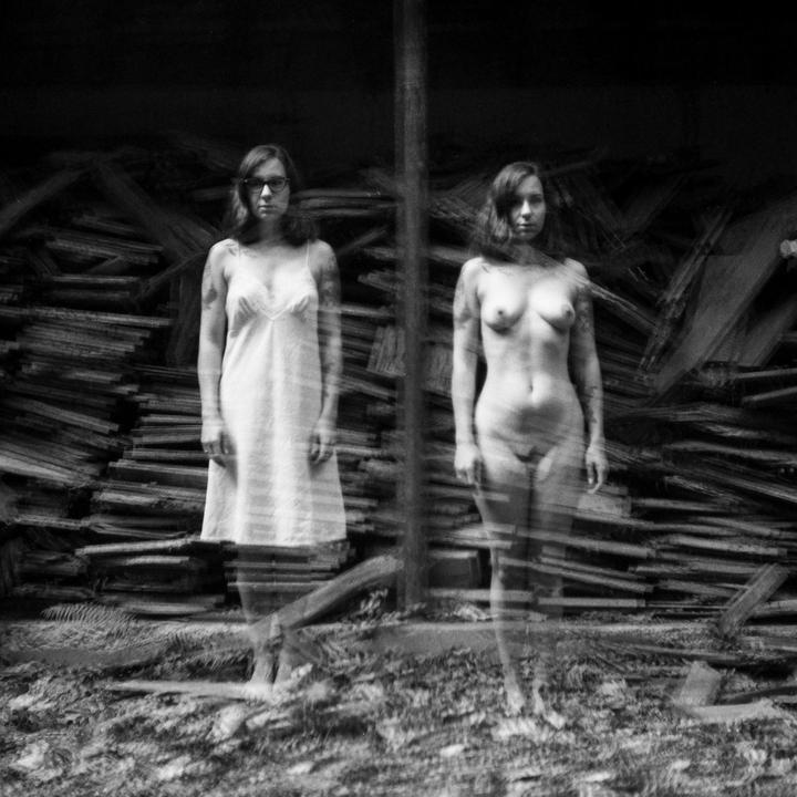 A woman in a slip and a nude woman stand side-by-side in front of a pile of wood.