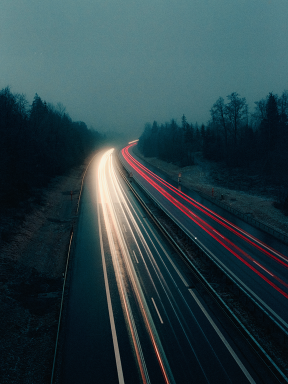 Light trails from vehicles illuminate a highway at night, flanked by dark trees under a hazy sky.
