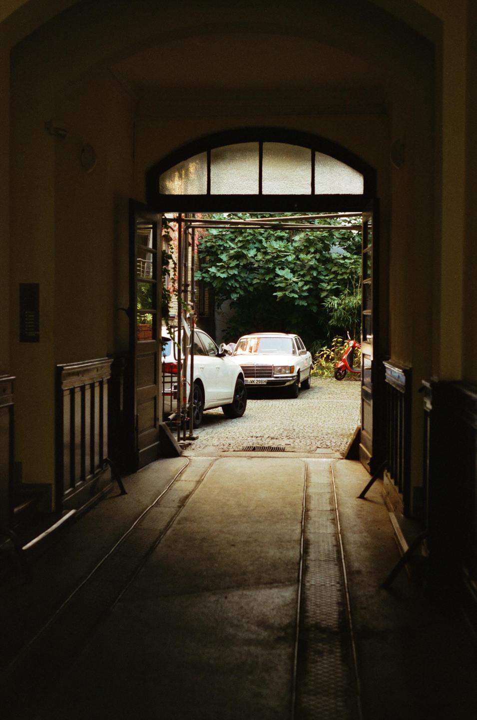 A dark hallway opens to a sunlit courtyard with two cars and a scooter.