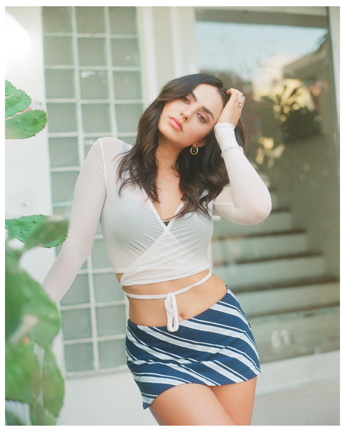 A woman with dark hair poses with her hand on her head, wearing a white long-sleeve top and a blue and white striped skirt, with a cactus in the foreground.