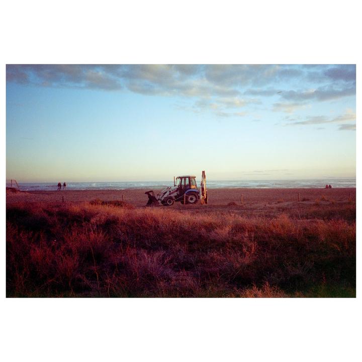 A blue and white tractor is parked on a beach with dry, reddish vegetation in the foreground and the ocean under a cloudy sky in the background.