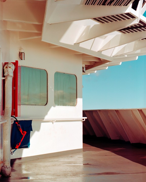 Sunlight casts shadows on the white walls and deck of a ferry, with a clear blue sky visible in the background.