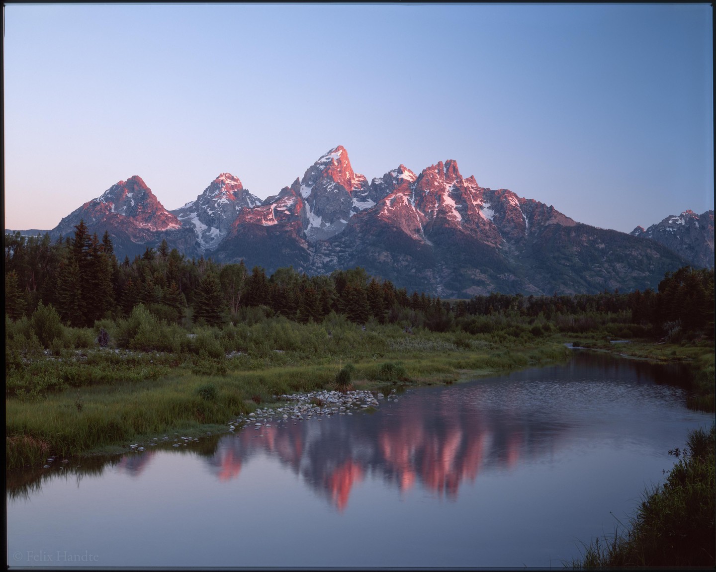 Snow-capped mountains are reflected in a river at sunrise, with a forest in the middle ground.