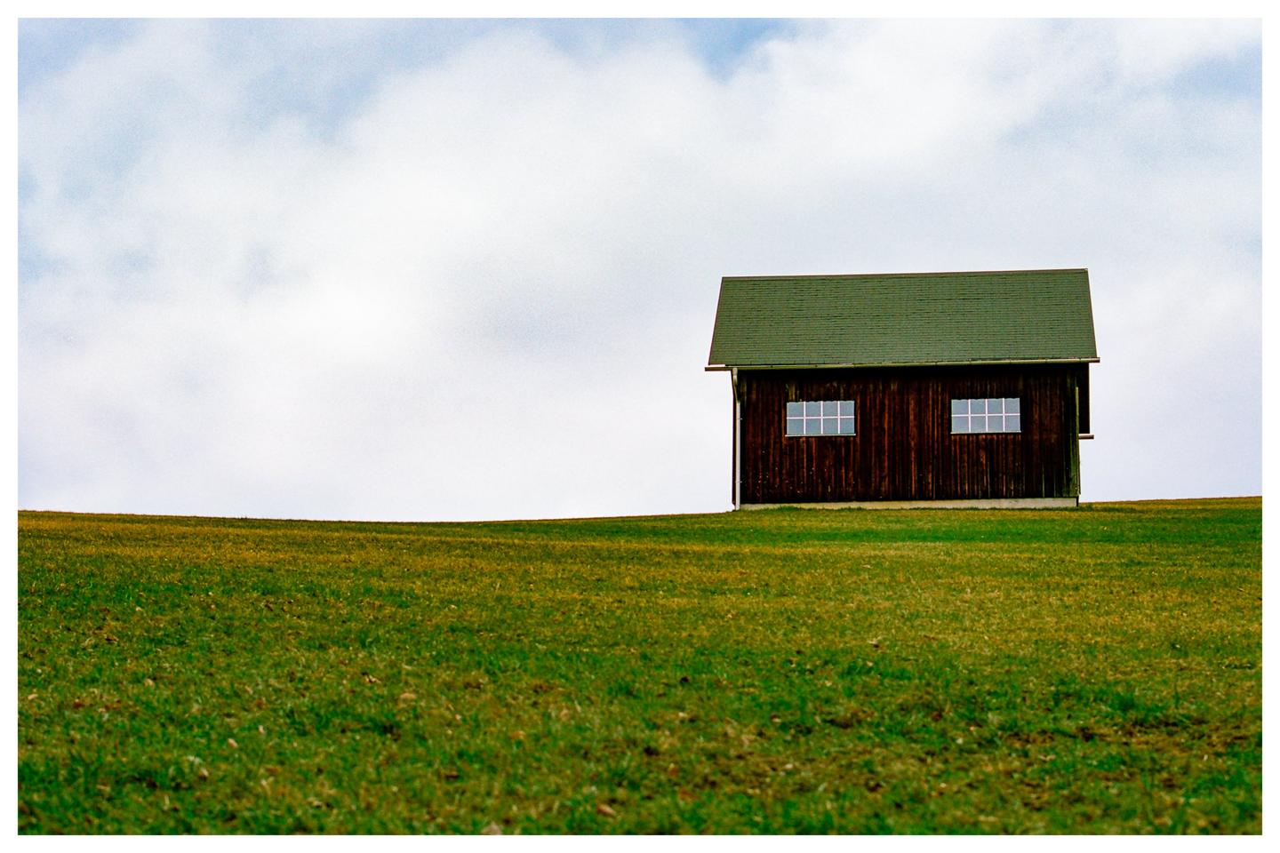 A small wooden house with a green roof sits on a grassy hill under a cloudy sky.