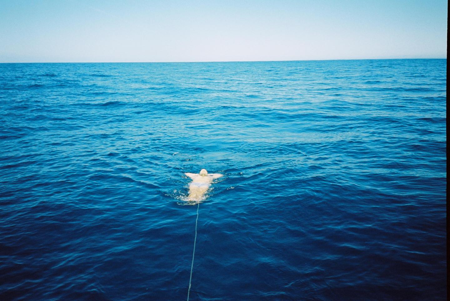 A person in a light-colored swimsuit floats on their back in the deep blue ocean, tethered by a rope.