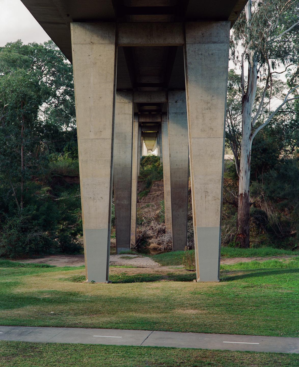 A concrete bridge extends over a grassy area with trees and a small stream.