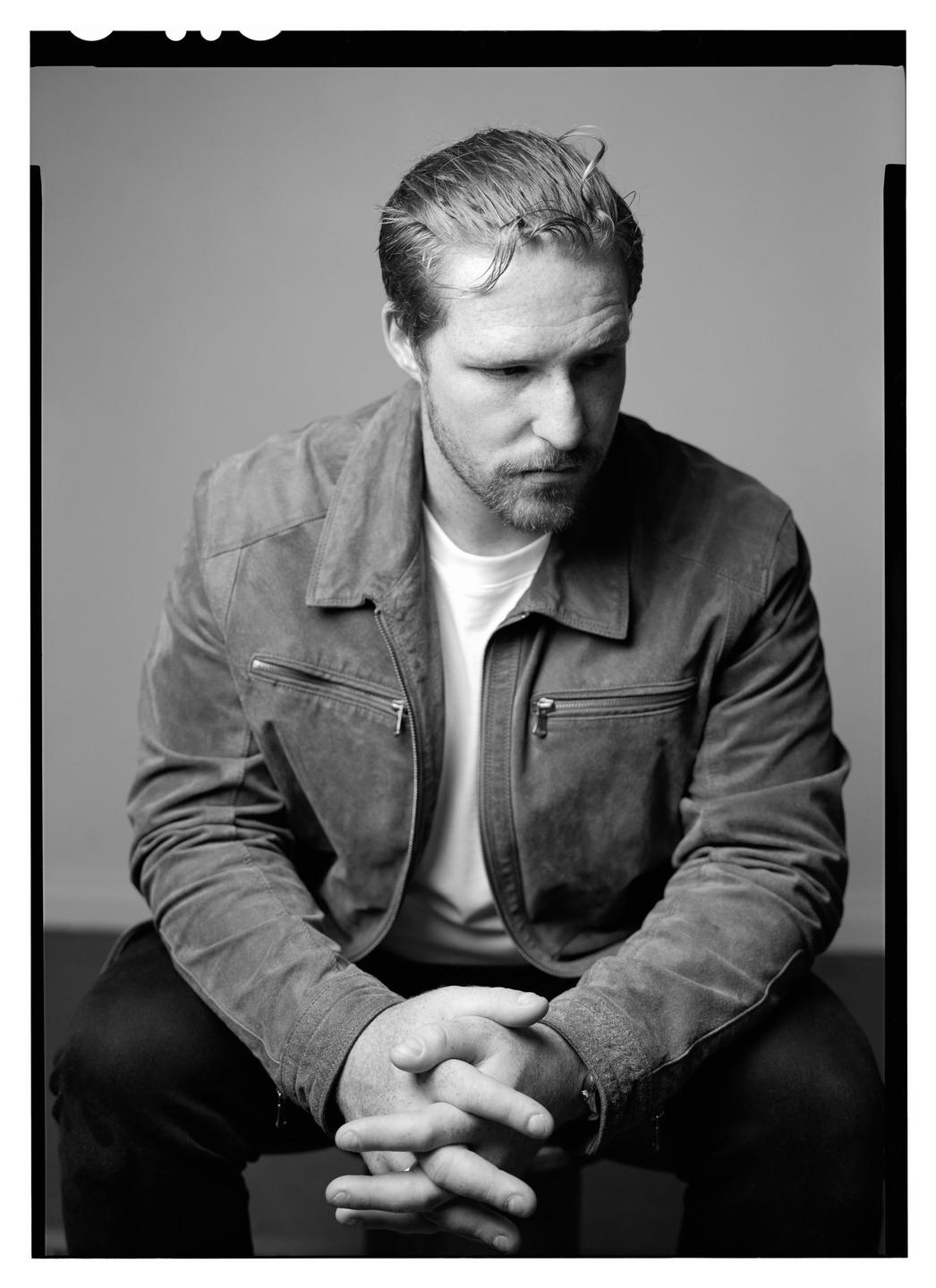 A man with a beard and slicked-back hair sits with his hands clasped, looking down.