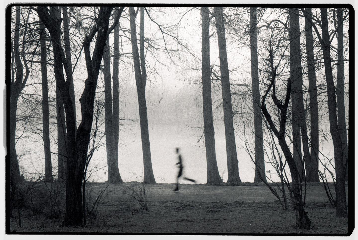 A blurry figure runs through a foggy forest with tall, bare trees.