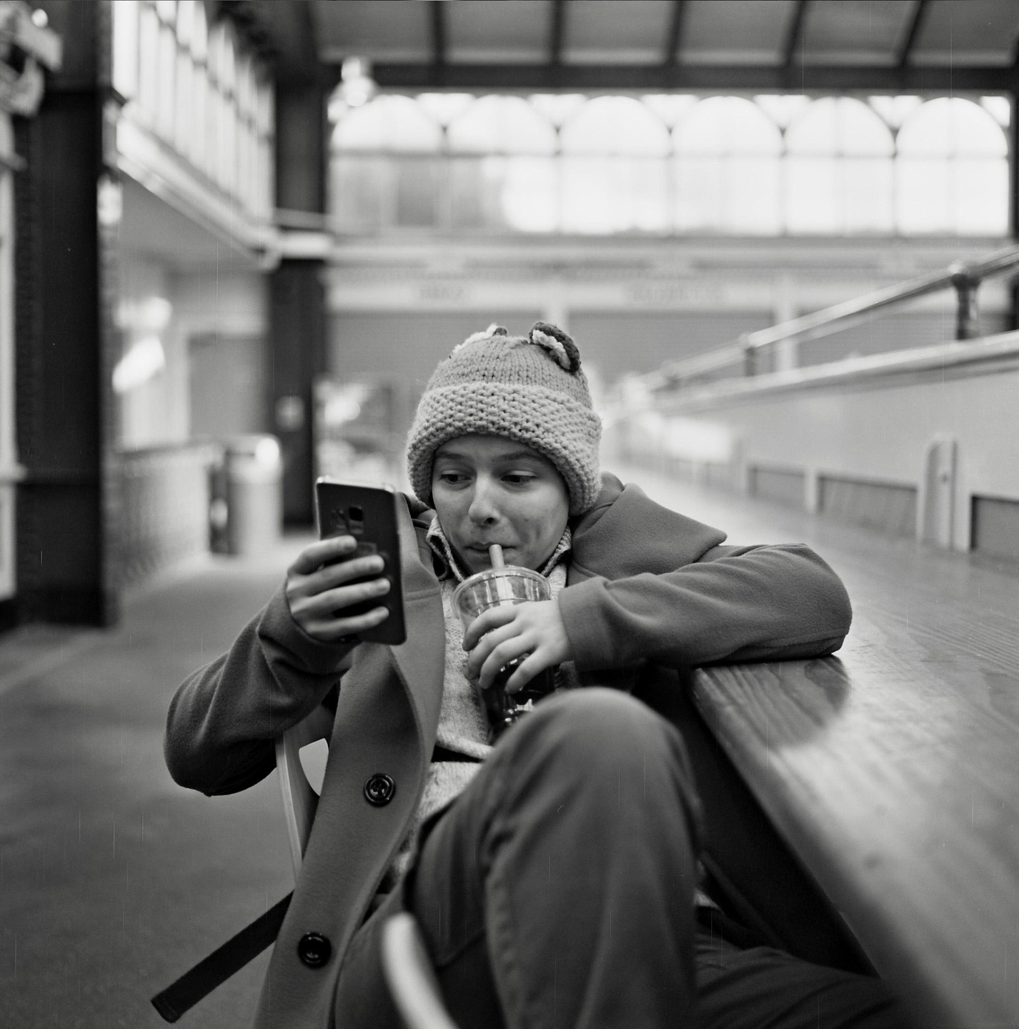 A young boy wearing a knit hat and coat sips a drink while looking at a phone.