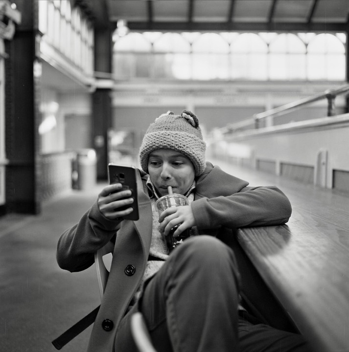 A young boy wearing a knit hat and coat sips a drink while looking at a phone.