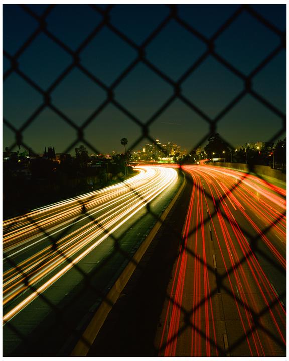 A highway at night is seen through a chain link fence, with light trails from vehicles on the road.