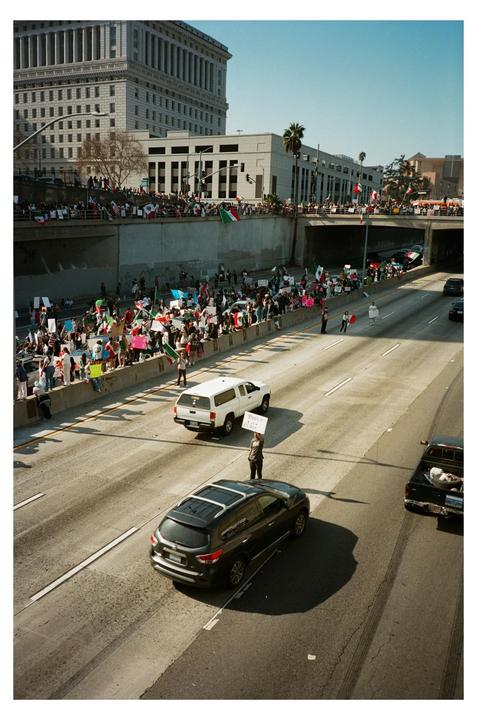 A large crowd of people with flags and signs are gathered on a bridge overlooking a highway with cars and a person holding a sign.