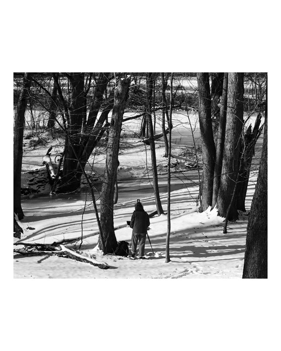 A person stands with a tripod in a snowy forest with bare trees and a frozen stream.