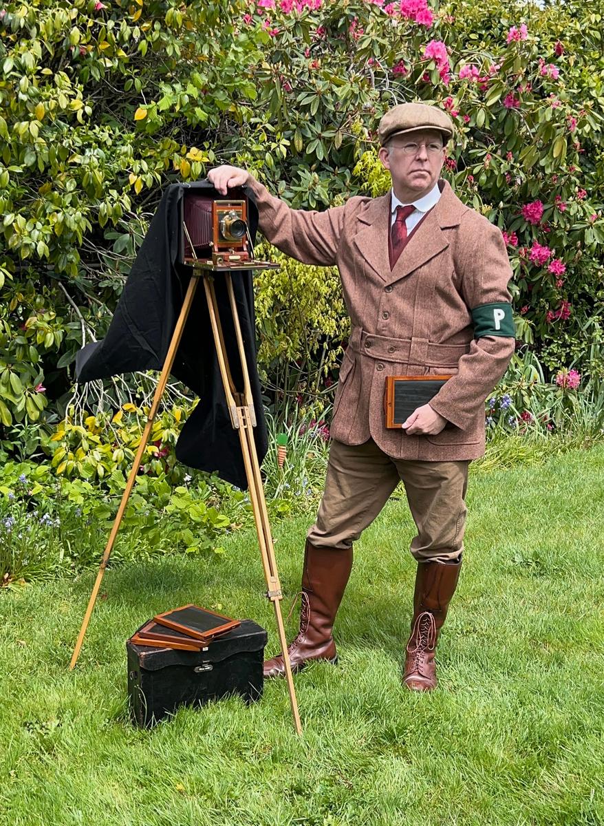 A man in period clothing stands next to a large format camera on a tripod in a garden.
