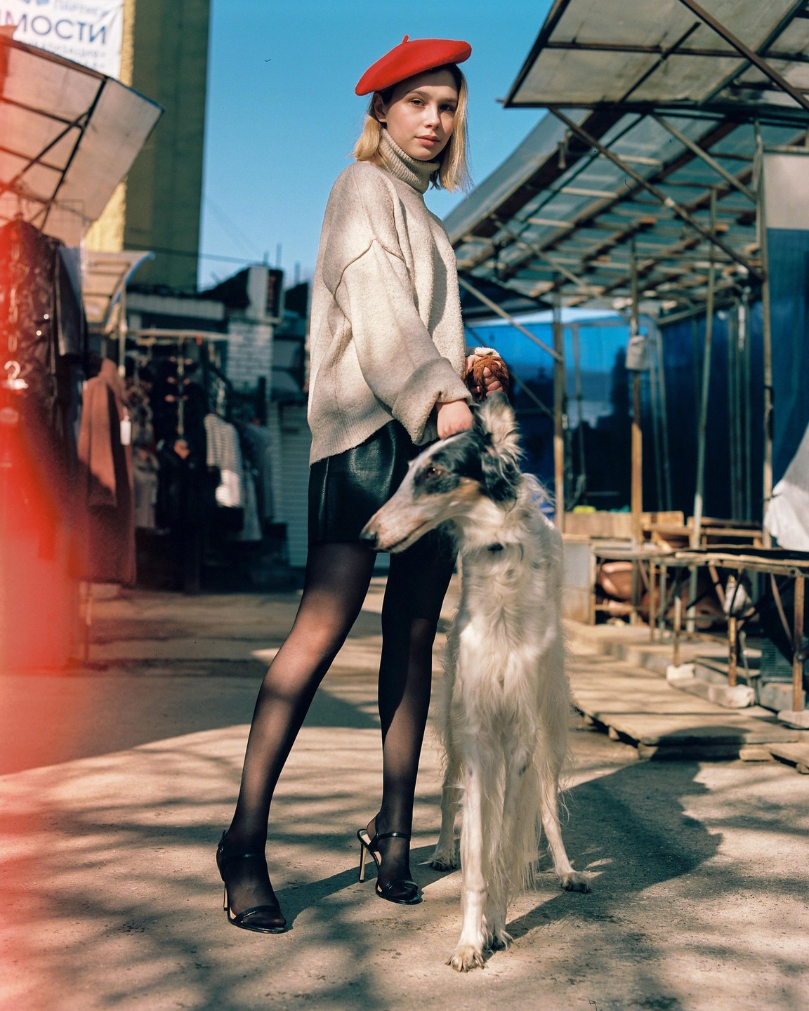 A woman in a red beret and a dog stand on a paved surface with market stalls in the background.