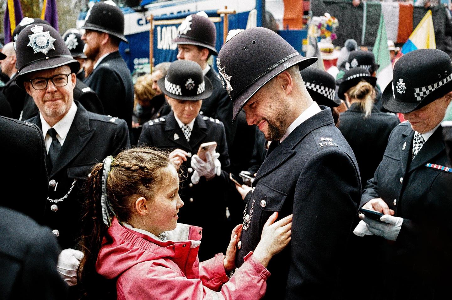 A girl in a pink jacket touches the uniform of a smiling police officer in a crowd of officers.