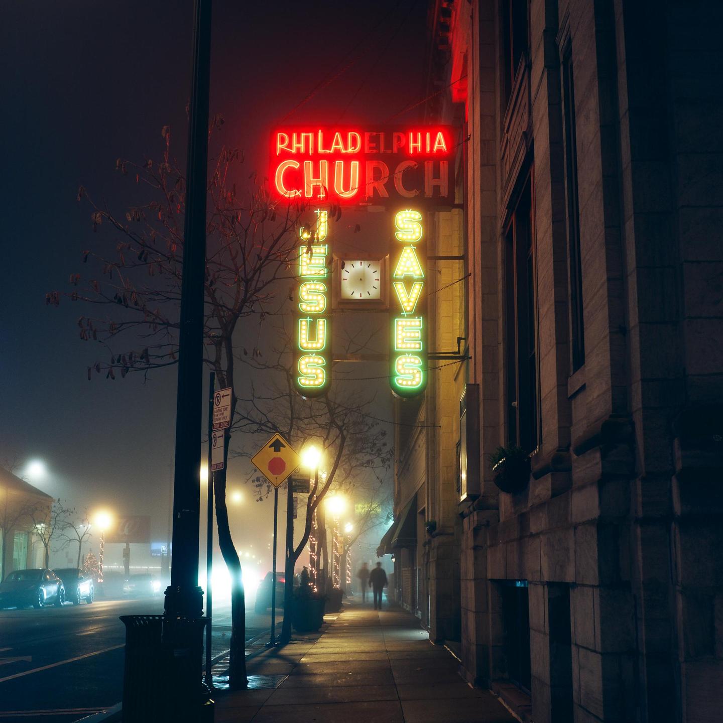 A neon sign for Philadelphia Church with the words "Jesus Saves" is illuminated on a foggy street at night.