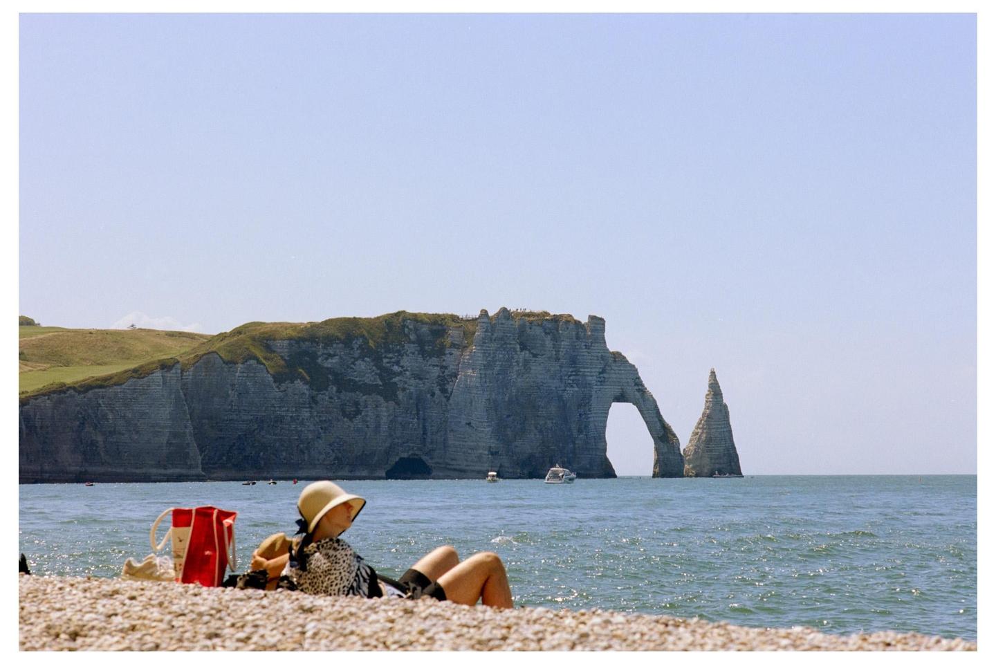 A woman wearing a sun hat relaxes on a pebble beach with cliffs and a natural arch in the background.