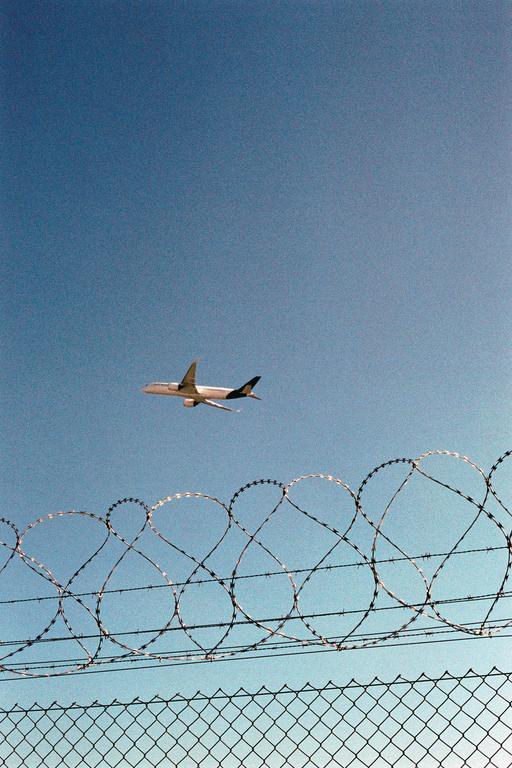 A passenger airplane flies in a clear blue sky above a chain-link fence topped with razor wire.
