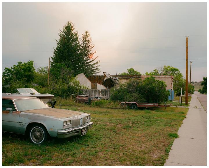 An old car is parked on a grassy lot next to a sidewalk with a dilapidated building and trees in the background.