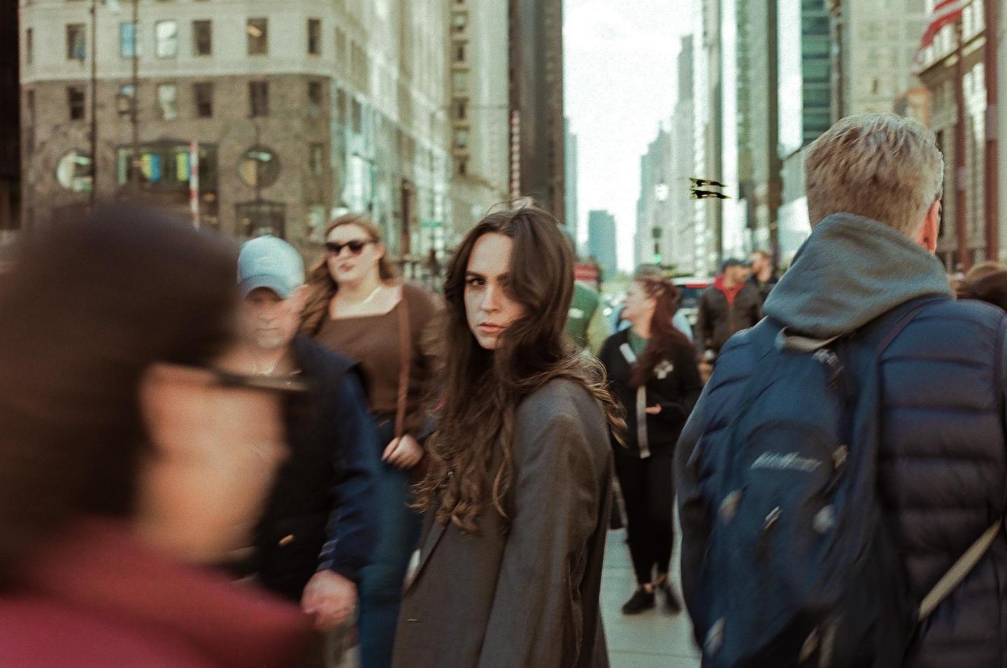A woman with long hair looks over her shoulder at the viewer while walking on a city street among other pedestrians and tall buildings.