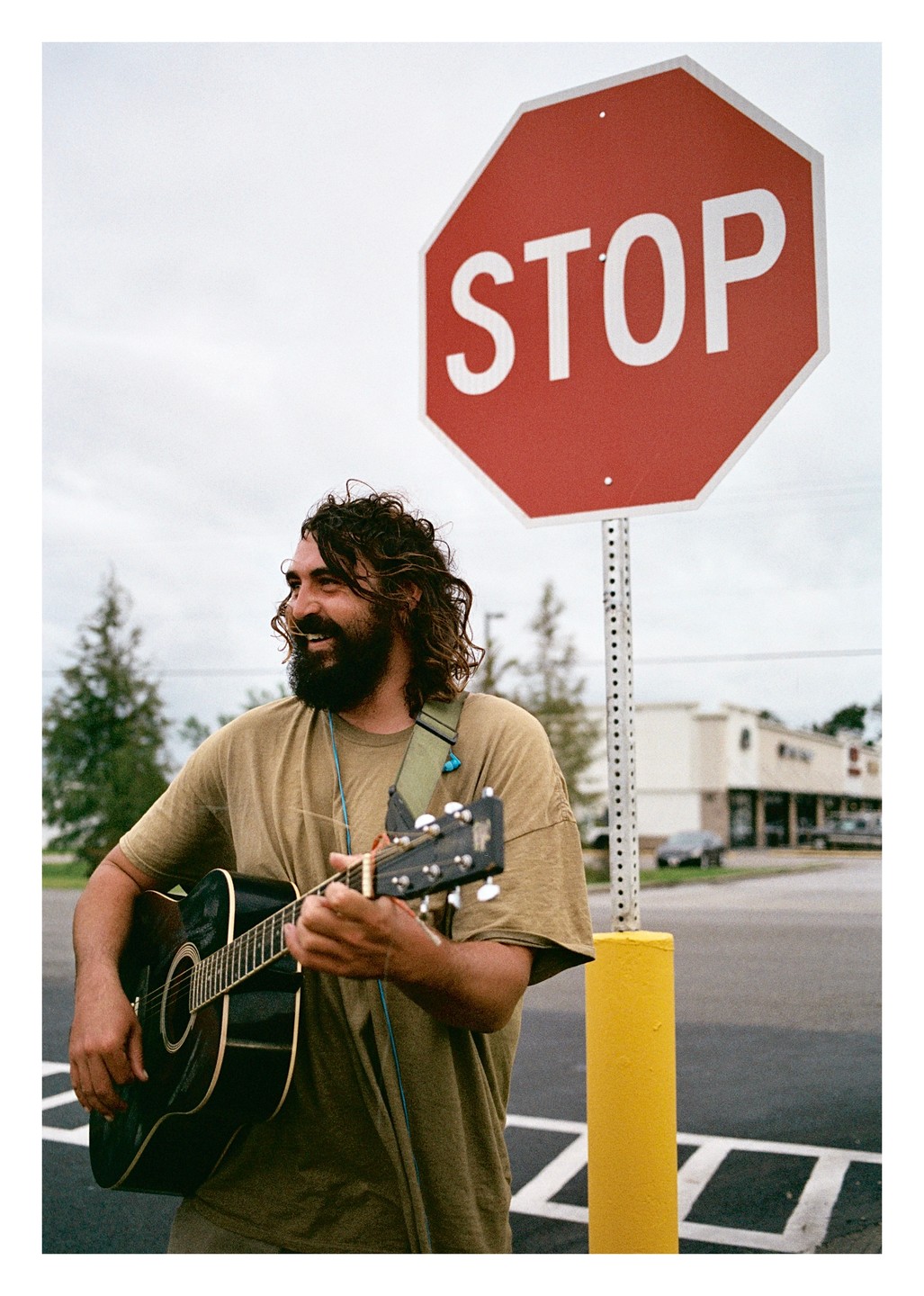 A smiling man with a beard and long hair plays an acoustic guitar next to a stop sign in a parking lot.