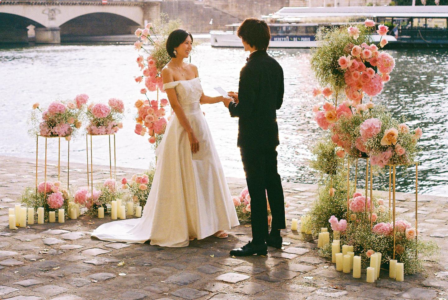 A couple stands by a river surrounded by flowers and candles, with a bridge and boat in the background.