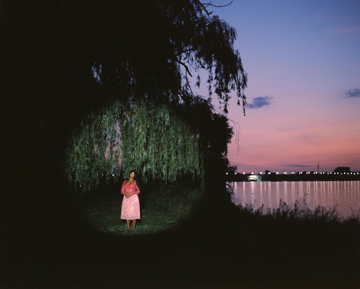 A woman stands in a circle of light under a weeping willow tree at dusk, with a body of water and lights in the background.