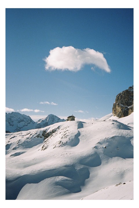 A small hut sits atop a snow-covered mountain with a single cloud in the blue sky.