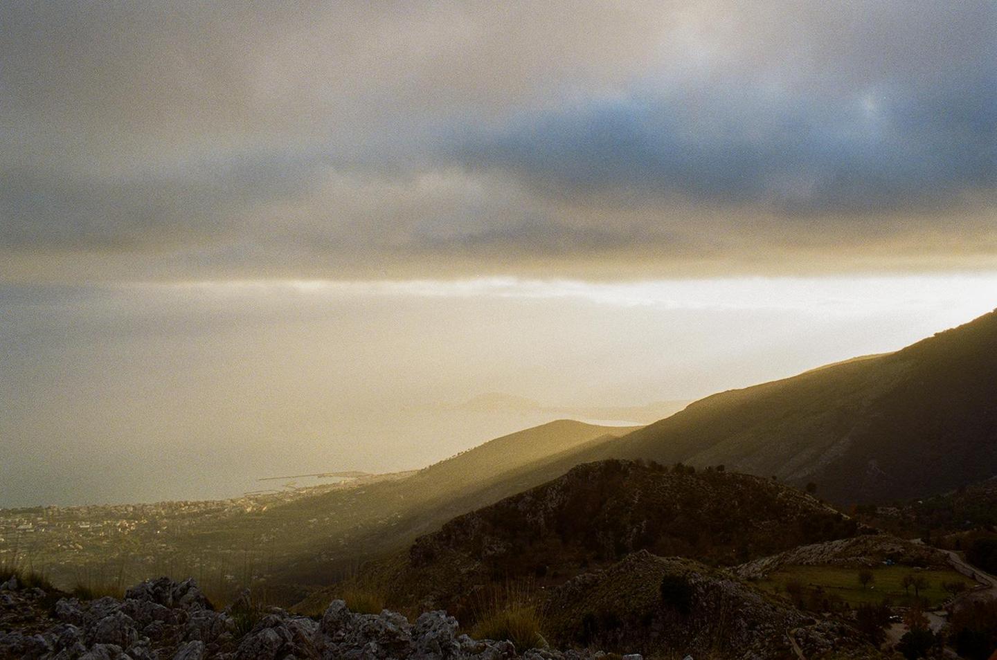 Sunlight breaks through clouds, illuminating a valley and distant town next to a body of water.