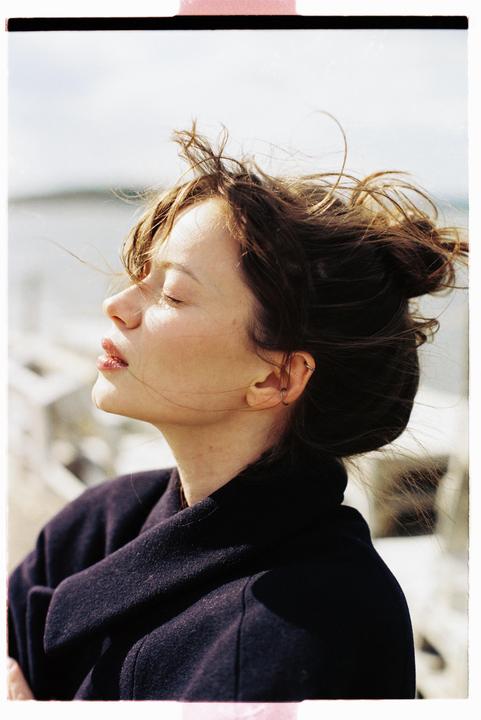 A woman with her eyes closed and hair blowing in the wind is illuminated by sunlight.