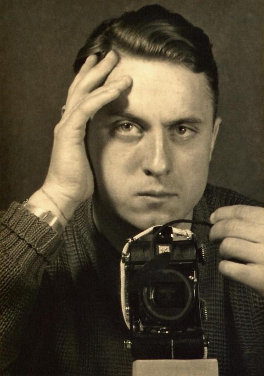 A man with his hand on his forehead looks directly at the viewer while holding a camera.