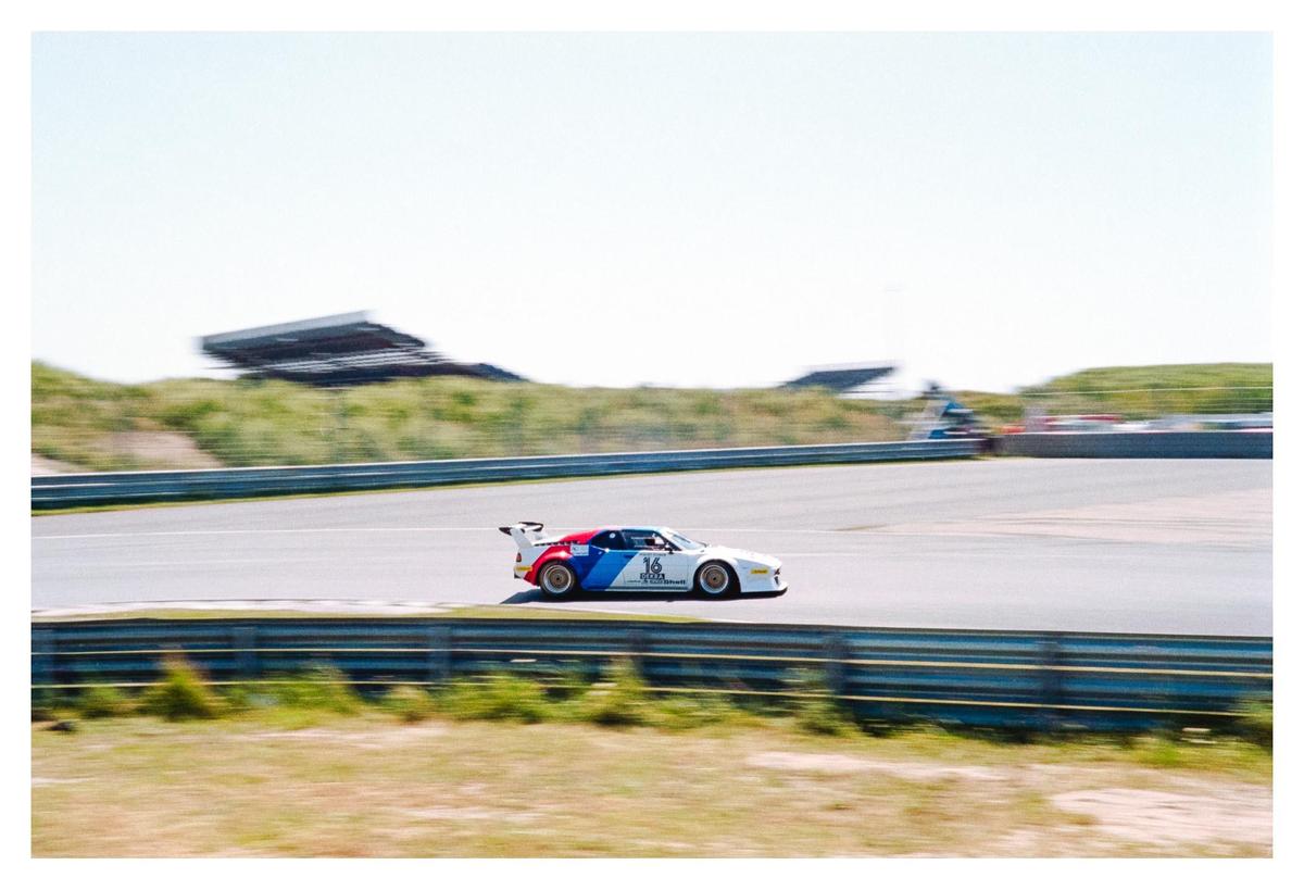 A white race car with red and blue stripes speeds around a track under a clear sky.
