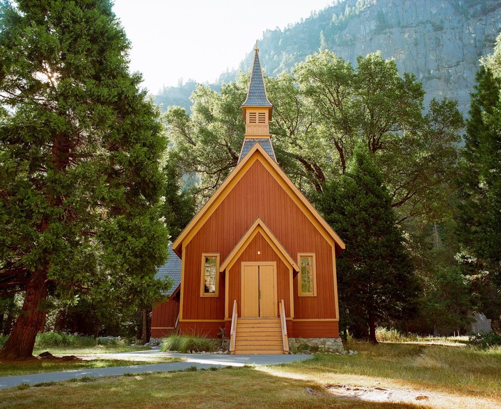 A small, rustic wooden church with a steeple stands among tall trees and a grassy area, with a mountain in the background.