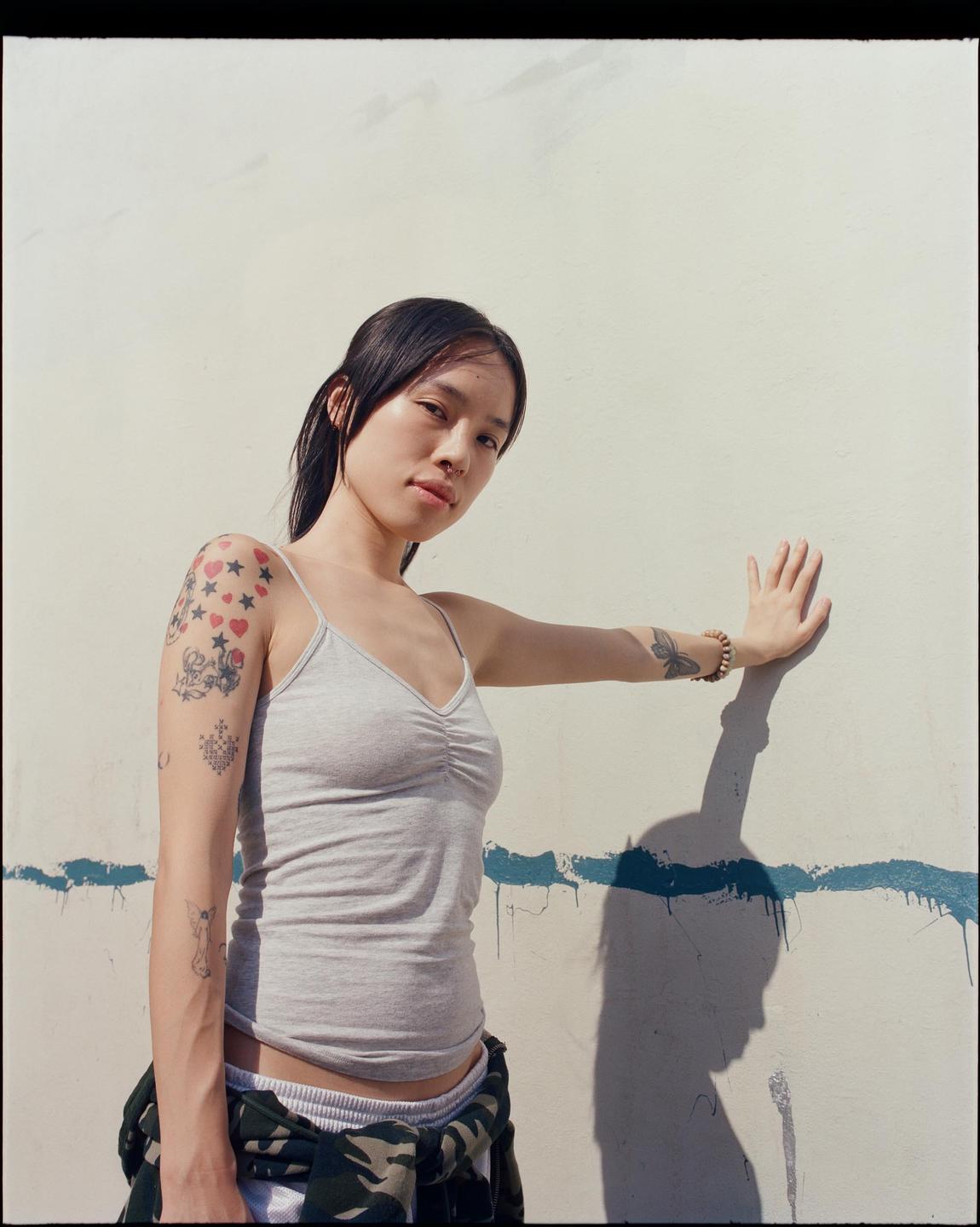 A woman with tattoos on her arm leans against a white wall with a blue stripe.
