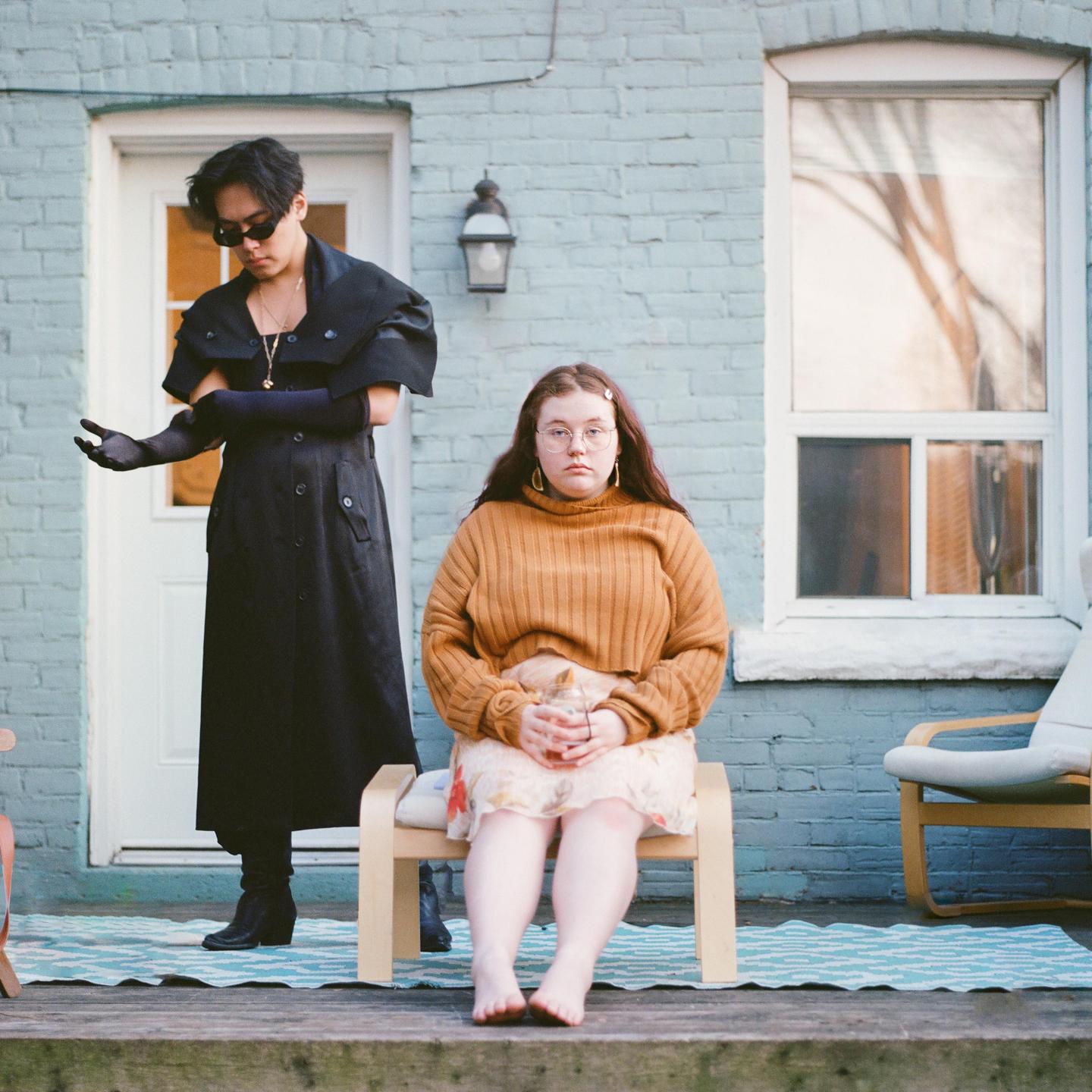 A woman sits on a wooden chair while a man stands beside her on a porch.