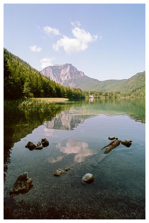 A clear lake with rocks and driftwood in the foreground reflects a mountain and sky with clouds, surrounded by a forest.