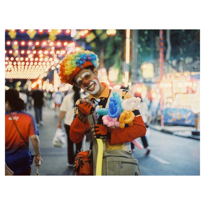 A street entertainer dressed as a clown smiles at the camera while holding balloon animals on a busy street at night.