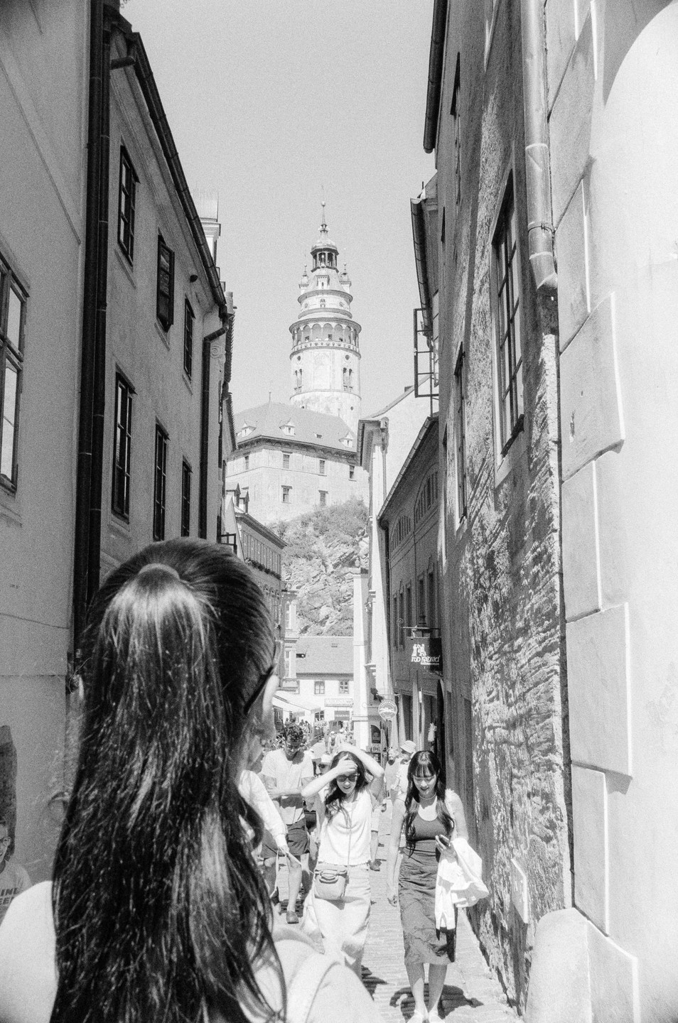 A woman with long hair and a ponytail walks down a narrow street toward a distant tower.