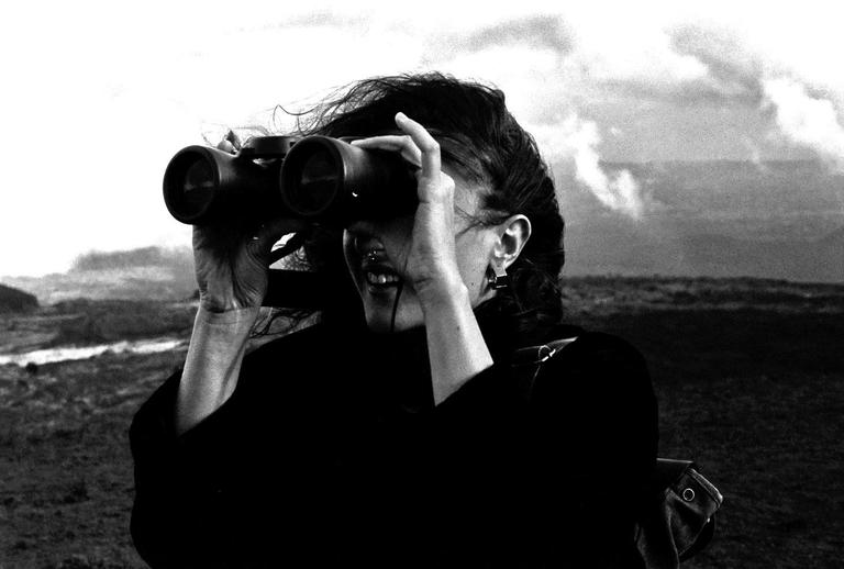 A woman with dark hair looks through binoculars with a wide smile, with a cloudy sky and a field in the background.