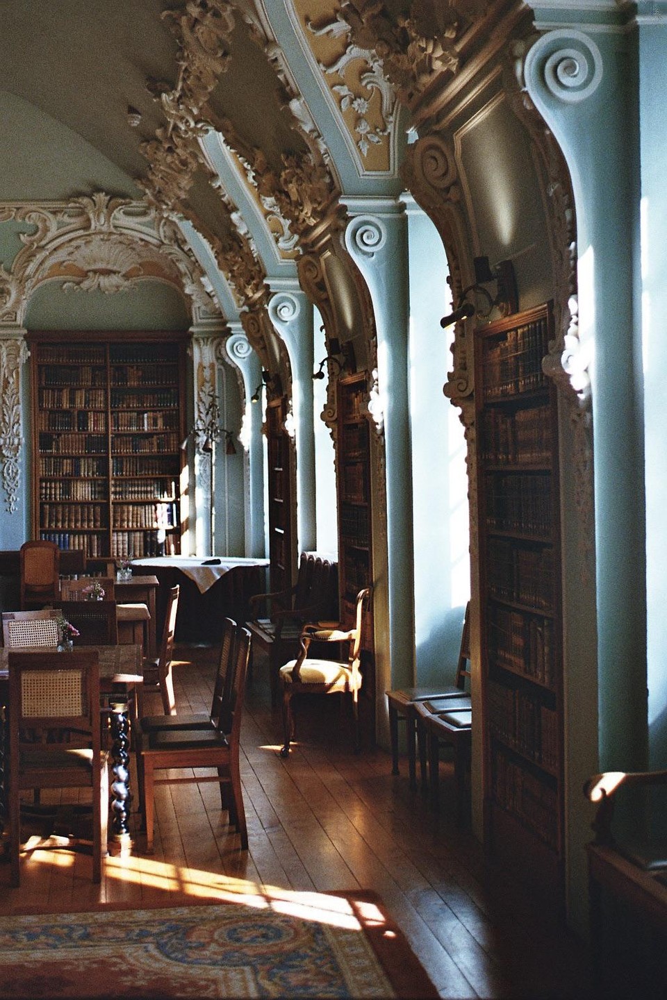 Sunlight streams into a rococo library with ornate columns, bookshelves, and wooden furniture.