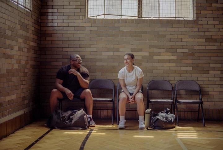 A man and a woman sit on chairs in a room with a brick wall and a wooden floor.