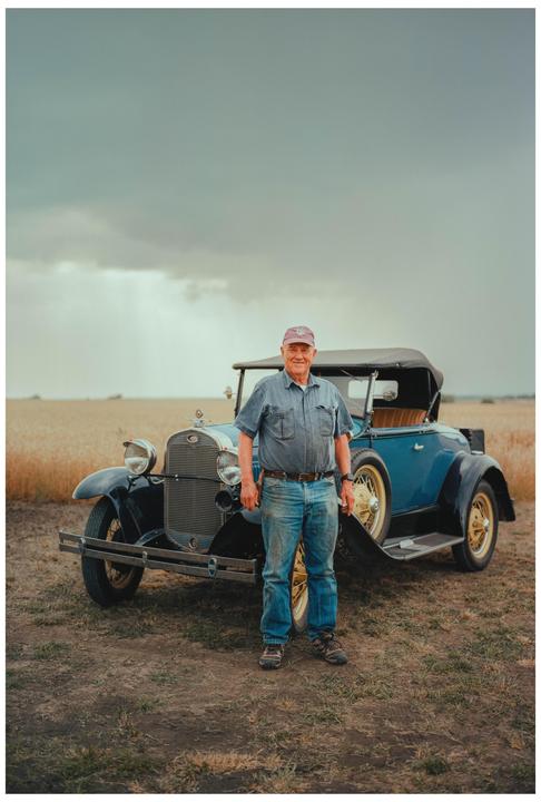 A man stands in front of a vintage blue car in a field under a cloudy sky.