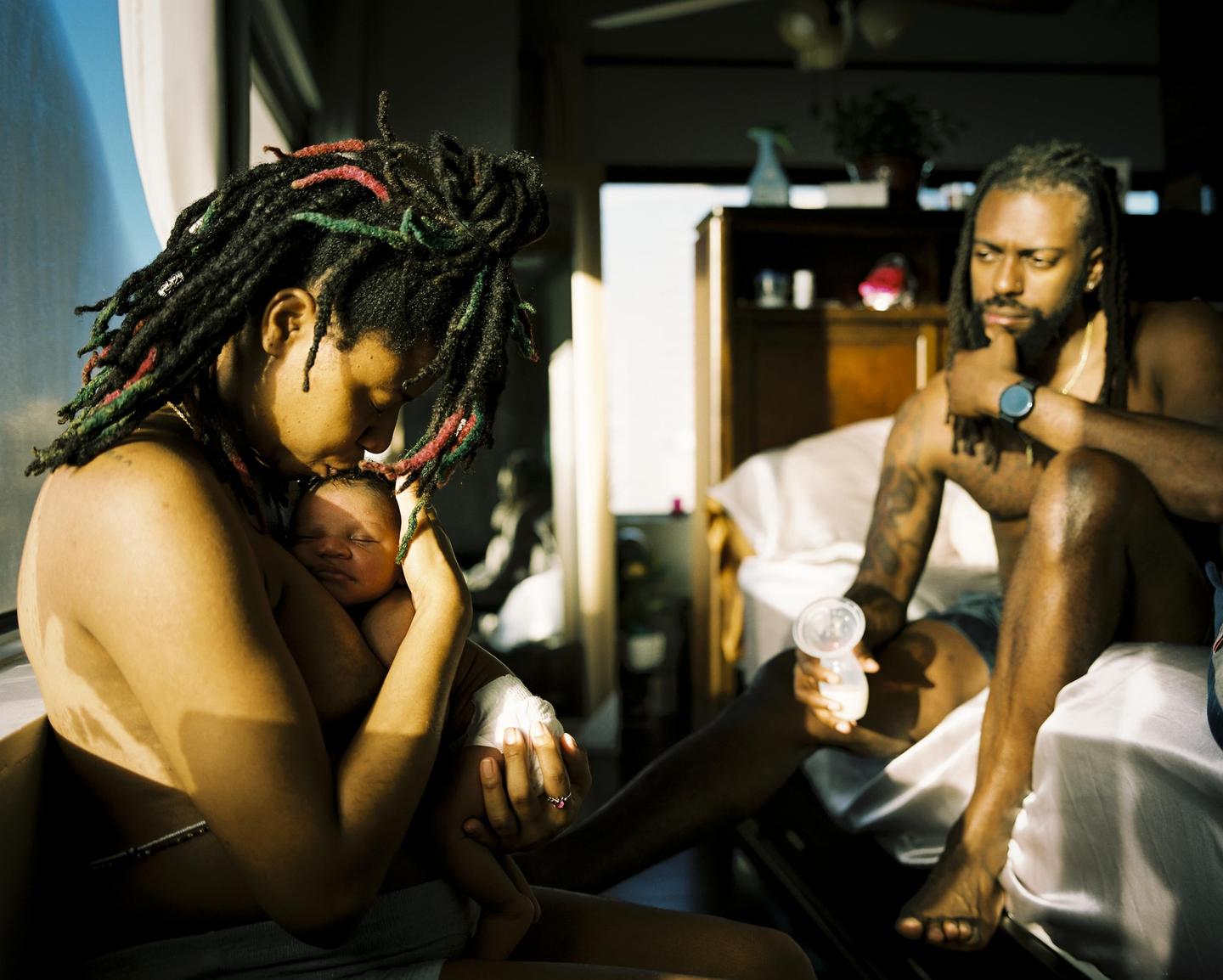 A woman with dreadlocks holds and kisses a sleeping baby while a man sits nearby holding a baby bottle.