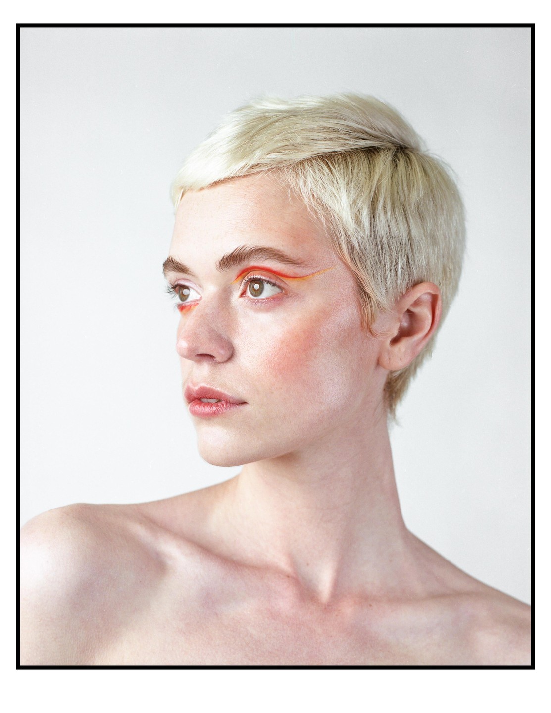 A woman with short blonde hair and orange eye makeup looks to the right.