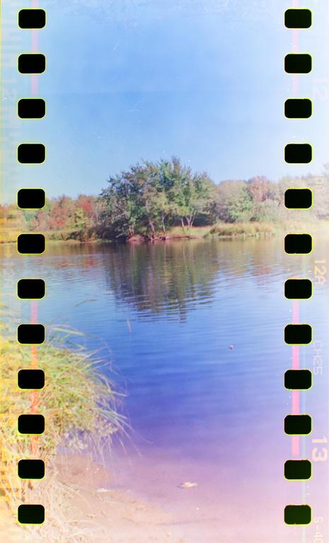 A body of water reflects the sky and trees on the opposite bank, with film sprockets visible on both sides of the image.