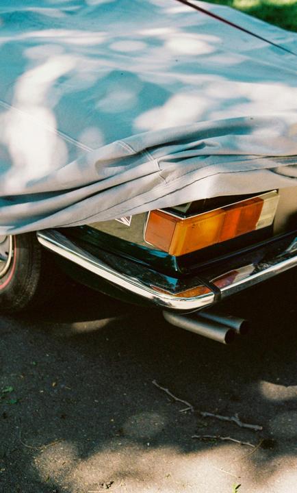 A car covered with a tarp is parked on asphalt, with its rear bumper, taillight, and exhaust pipes visible.