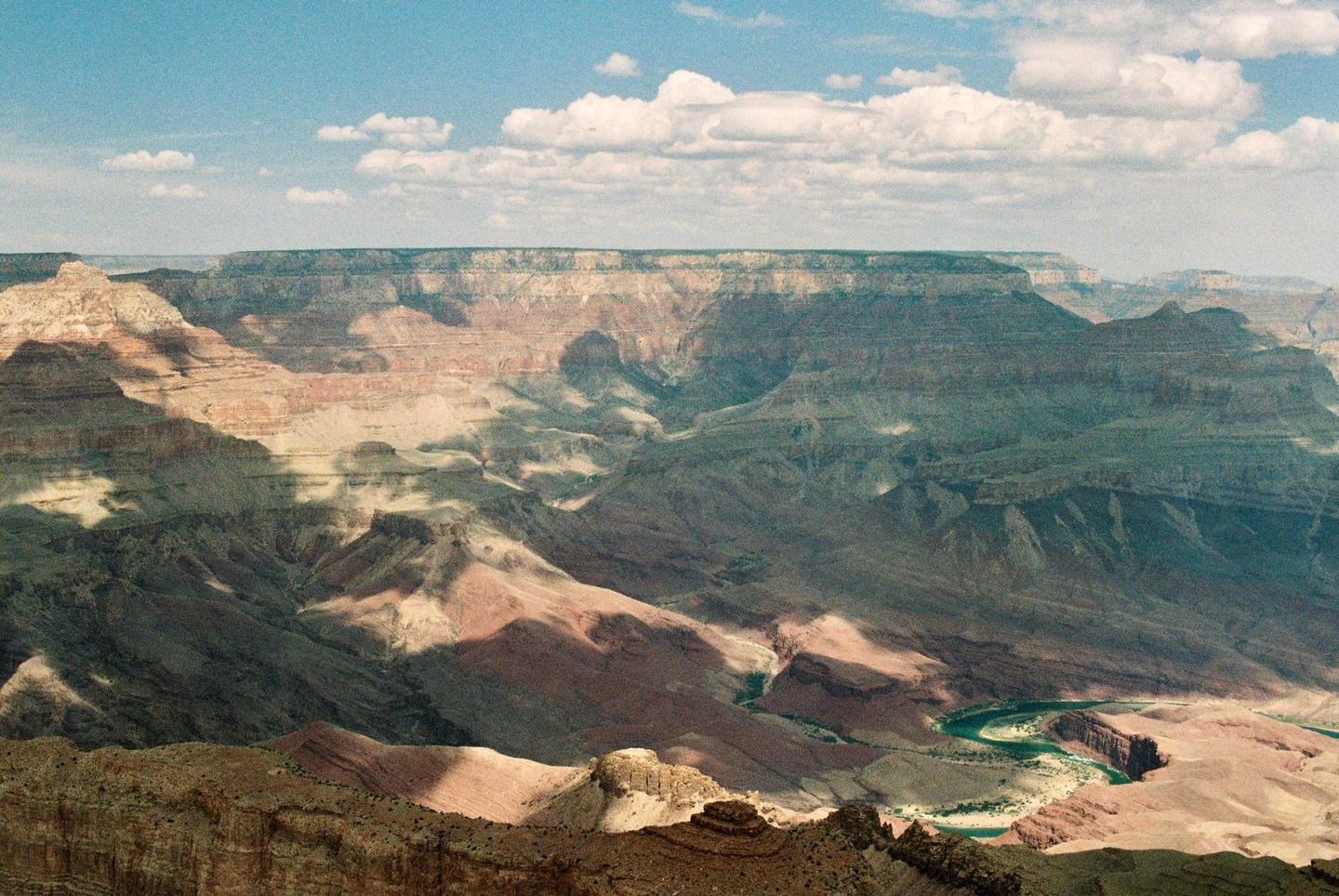 A wide view of the Grand Canyon shows a river winding through the bottom and clouds casting shadows on the canyon walls.