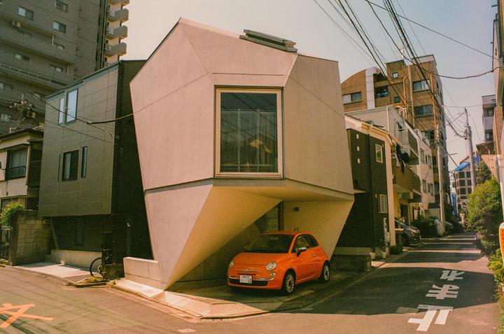 An orange car is parked under a modern, angular building on a street in Tokyo.